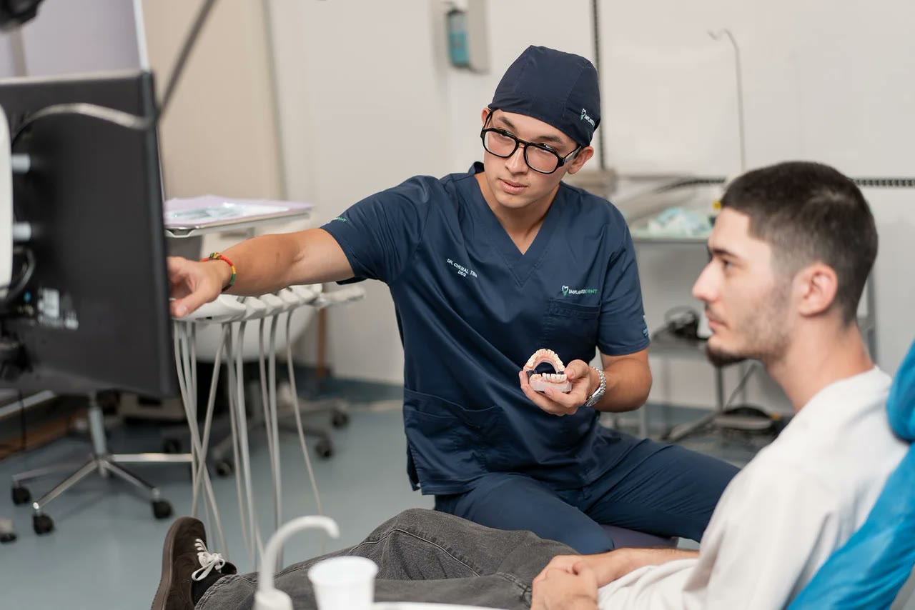 Medic implantolog explicând planul de tratament unui pacient, folosind un model dentar și imaginile afișate pe monitor