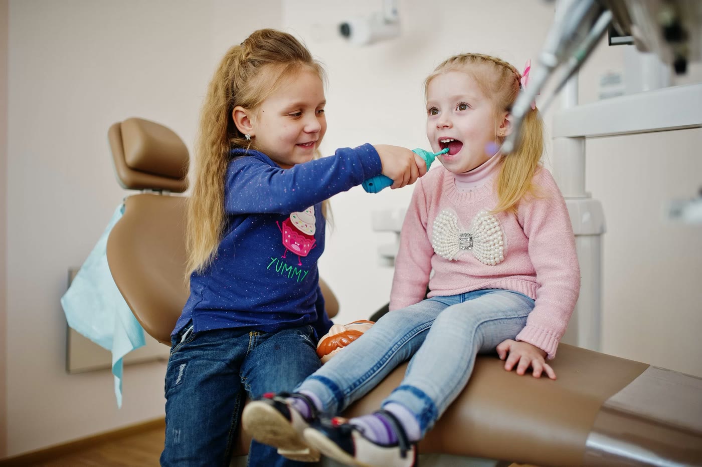 Două fetițe se joacă „de-a dentistul” pe scaunul stomatologic, una periind dinții celeilalte