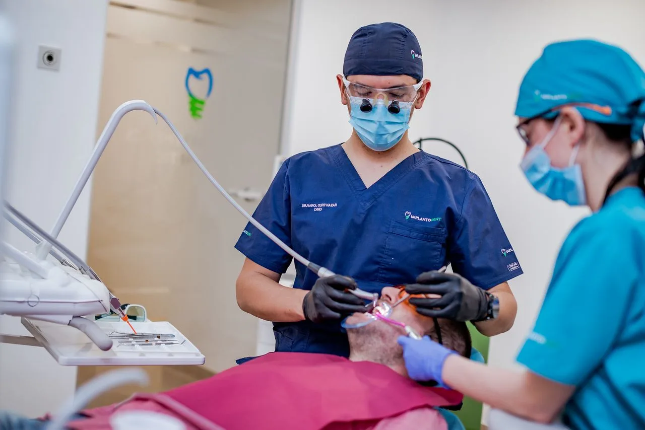 Medic Implantodent lucrând asupra unui pacient, asistat, în cabinet